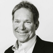 Fotografía en blanco y negro de un hombre sonriente con traje y camisa con cuello sobre un fondo liso.